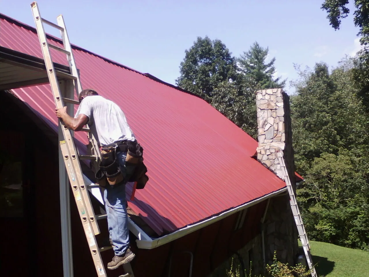 Roofer on a ladder beside a red metal roof and stone chimney — Soffit & Fascia Repair by Roofing Brothers in Mineral Springs