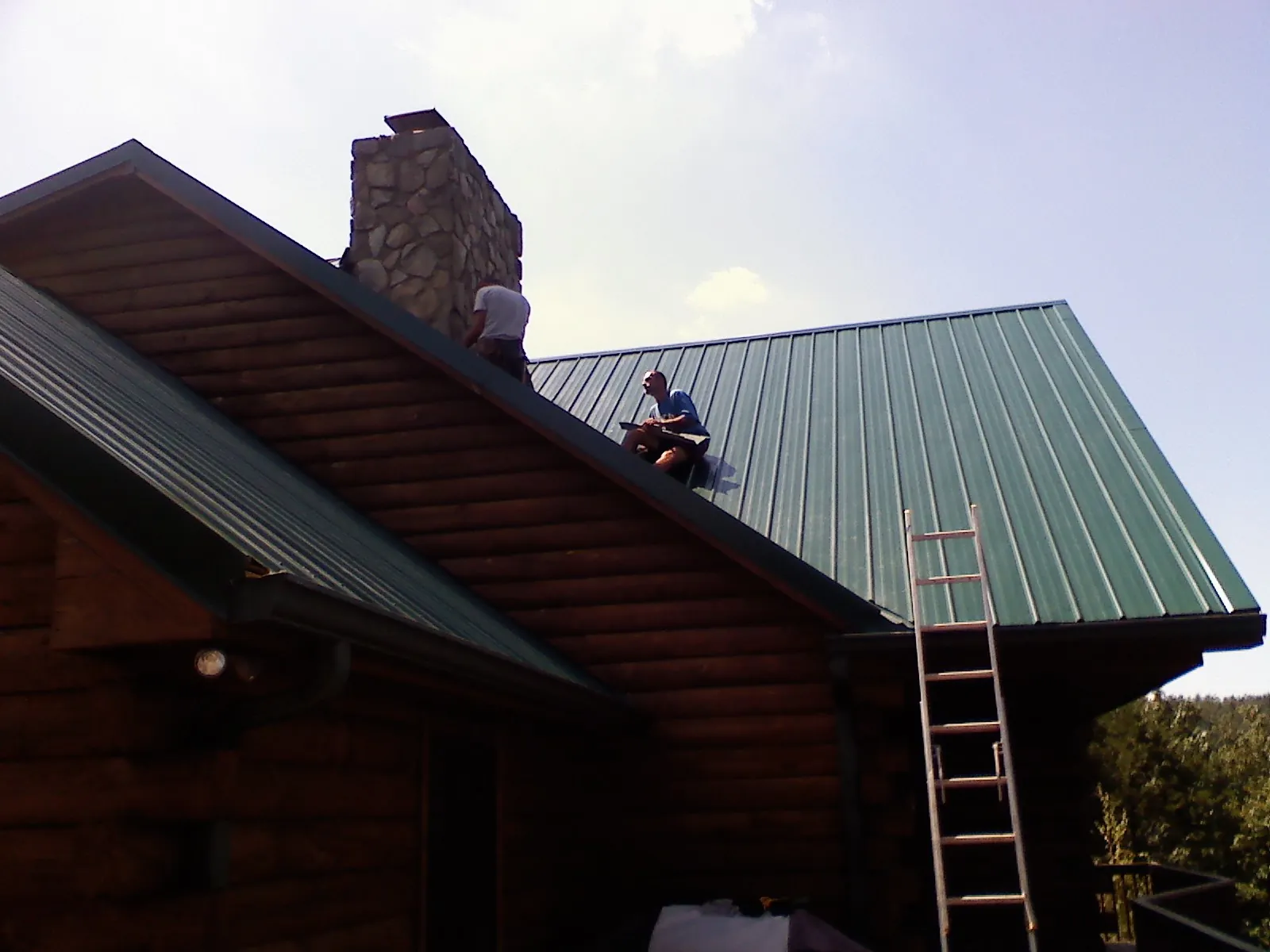 Roofers installing green metal panels beside a stone chimney on a log home in Mineral Springs